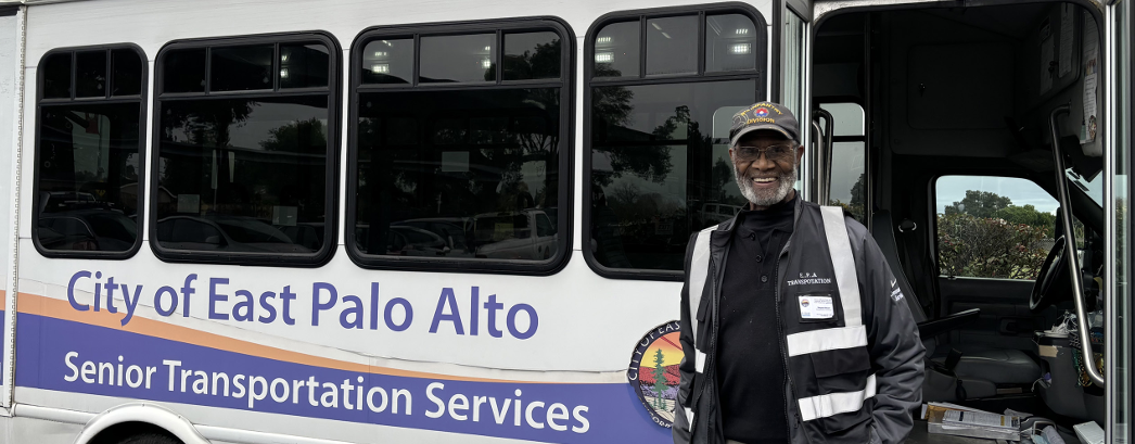 an older adult, standing and smiling, outside the door of a City of East Palo Alto bus.