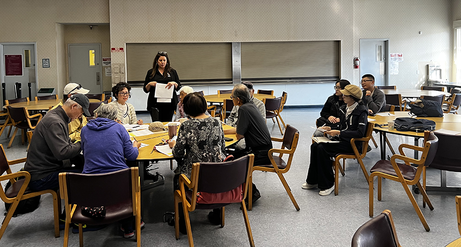 group of older Asian adults sitting at round tables, looking engrossed in reading documents, guided by a counselor.